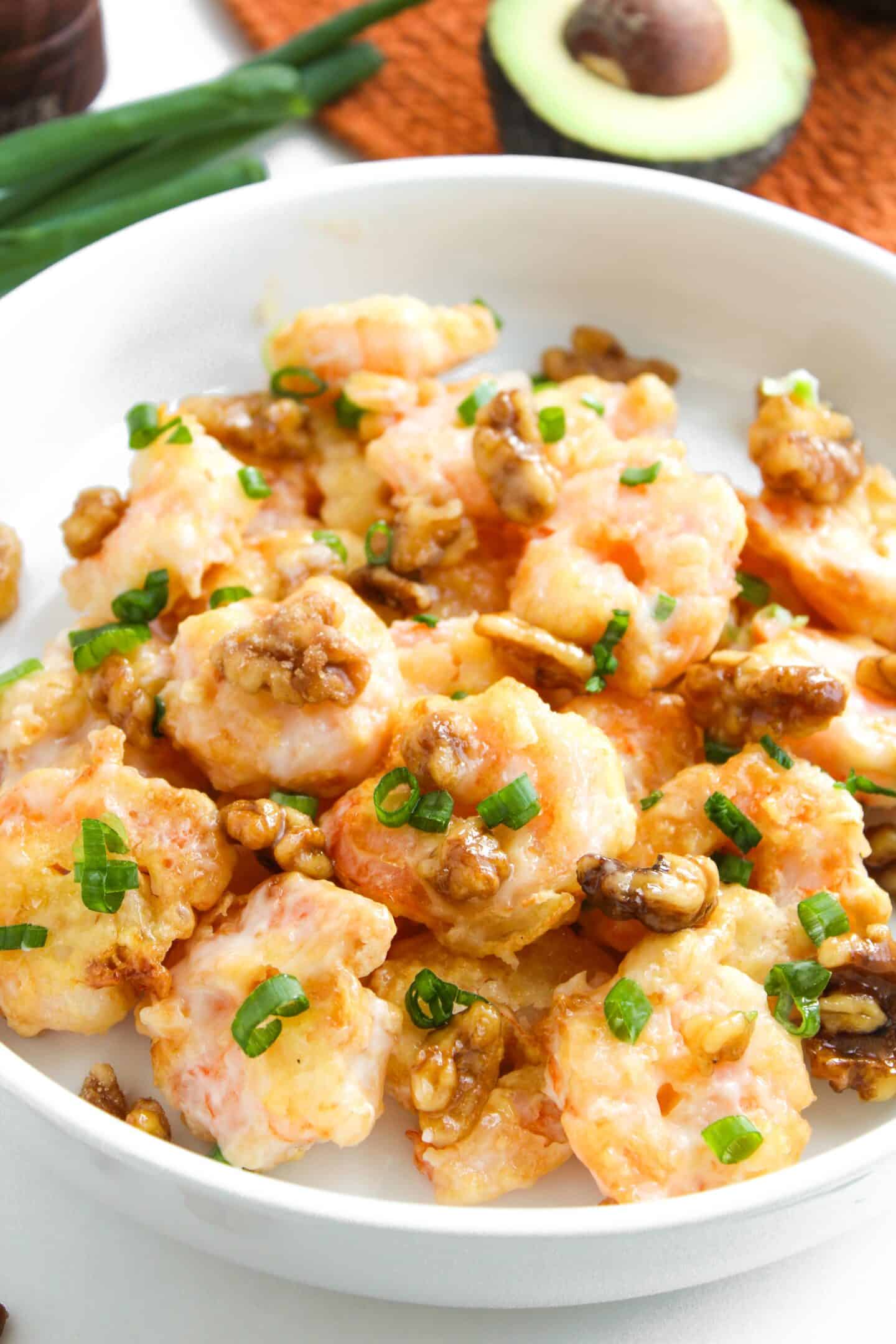 A white bowl filled with creamy Honey Walnut Shrimp topped with chopped green onions and walnuts, with an avocado half and green onions in the background.