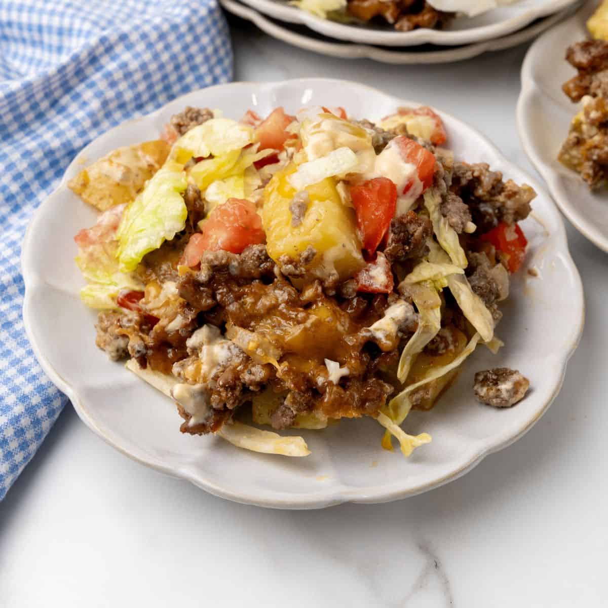 A plate of Big Mac-inspired cheeseburger casserole with ground beef, lettuce, tomatoes, and melted cheese sits invitingly on a white plate. A blue checkered cloth is casually placed nearby.