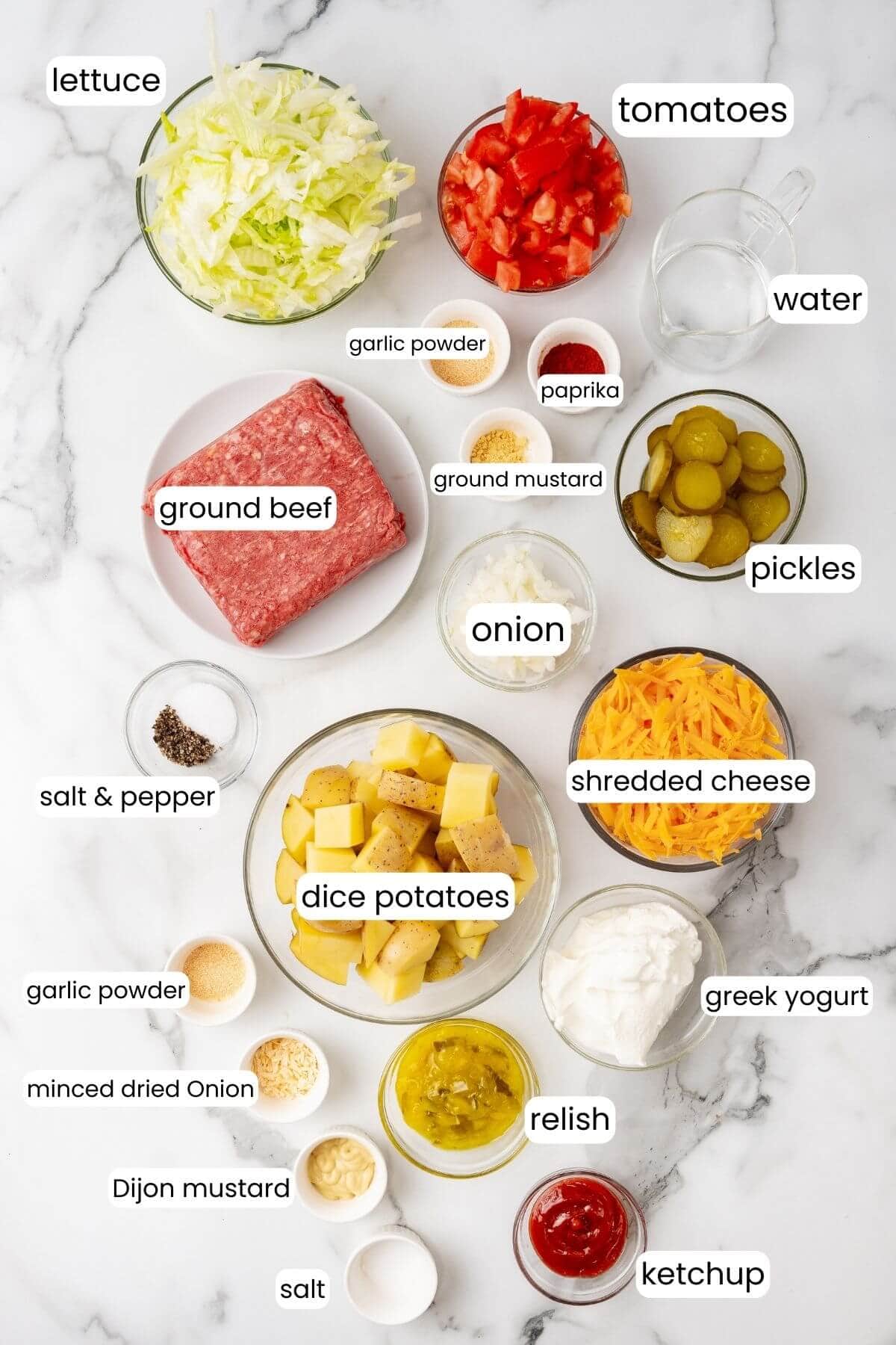 Ingredients arranged on a marble surface for a Big Mac-inspired casserole recipe, including ground beef, lettuce, tomatoes, diced potatoes, pickles, shredded cheese, seasonings, water, Greek yogurt, relish, and ketchup.