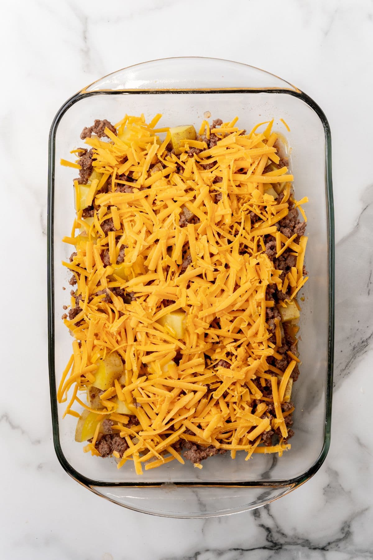 A hearty casserole brimming with ground meat and sliced potatoes, all crowned with a generous layer of shredded cheddar cheese, rests invitingly on the marble surface—a comfort food recipe that's sure to delight.