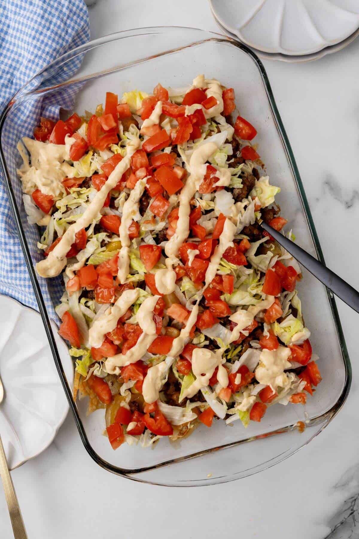 A glass dish showcases a vibrant, layered salad reminiscent of a Big Mac, with shredded lettuce and diced tomatoes drizzled in creamy dressing. A serving spoon rests inside, ready to serve. Nearby, a blue-and-white checked cloth adds a touch of charm.