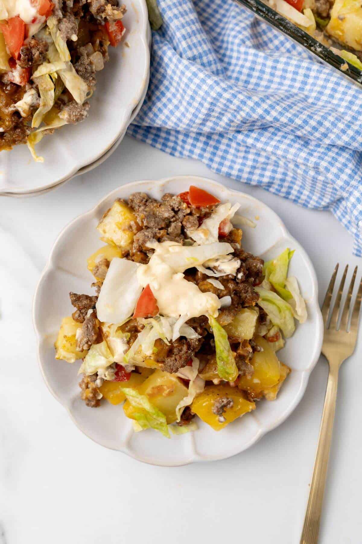 A delightful plate of taco casserole, reminiscent of a Big Mac. It features ground meat, potato chunks, crisp lettuce, juicy tomatoes, and creamy cheese sauce. A fork rests beside it on a checkered cloth backdrop.