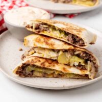 Three stacked quesadilla wedges filled with beef, cheese, onions, and pickles on a plate resemble a fun twist on the classic Big Mac, with a small bowl of creamy dip in the background.