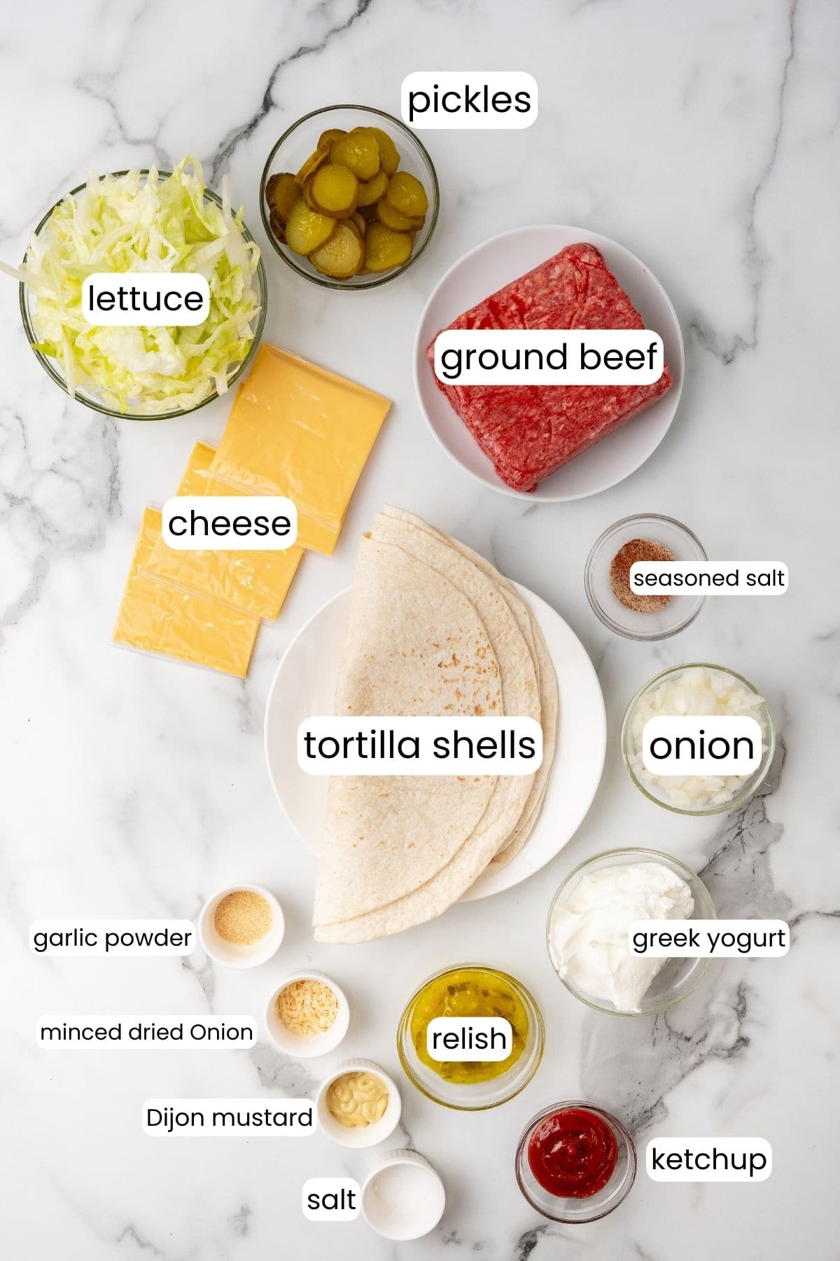 Ingredients for Big Mac-inspired cheeseburger wraps on a marble surface: tortilla shells, cheese, ground beef, lettuce, pickles, onion, relish, Greek yogurt, ketchup, mustard, garlic powder, seasoned salt. Indulge in a fusion of flavors with every bite!.