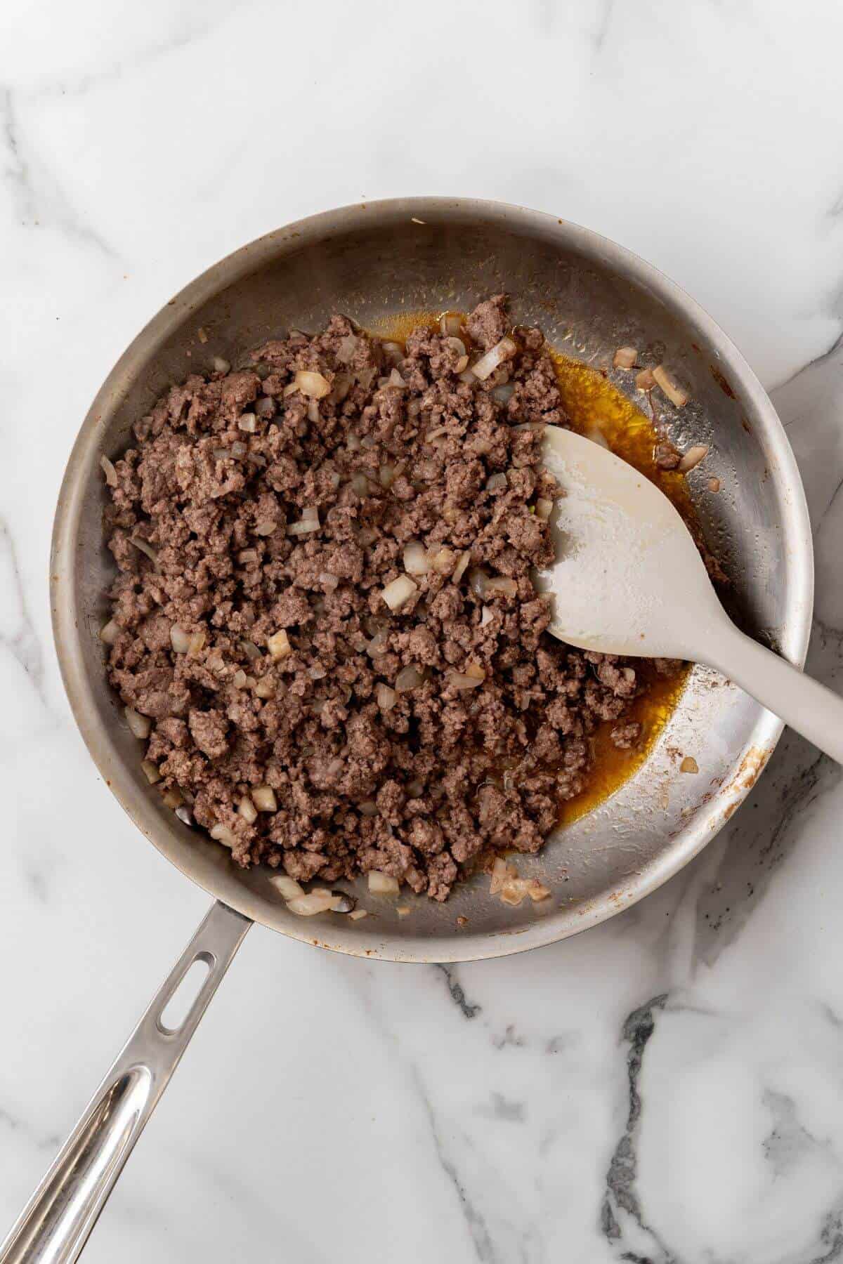 Ground beef with diced onions being cooked in a stainless steel pan on a marble countertop, stirred with a spatula, reminiscent of preparing the savory filling for a quesadilla.
