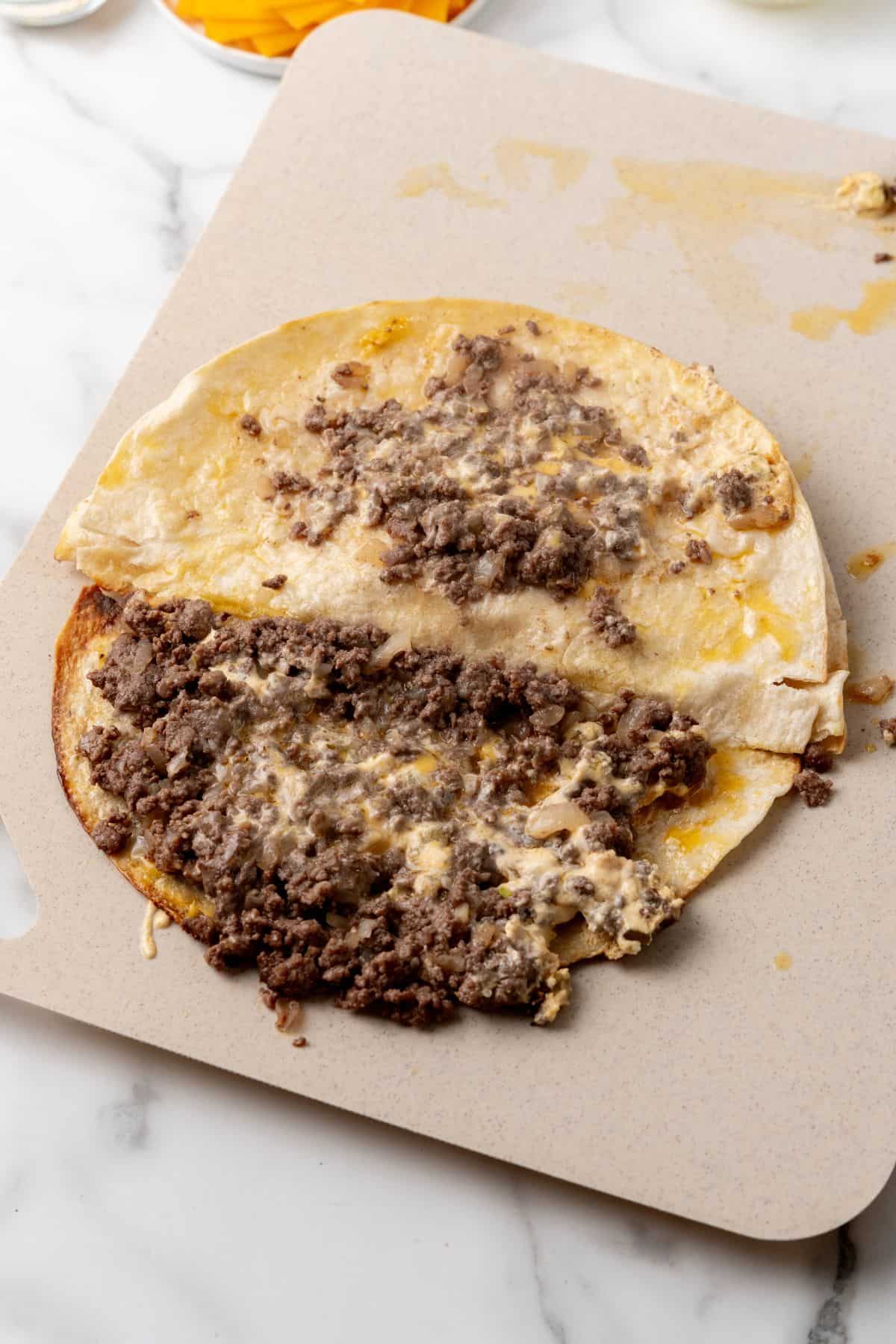 A quesadilla on a cutting board, folded in half like a savory version of a Big Mac, with cooked ground beef spilling out from the inside.