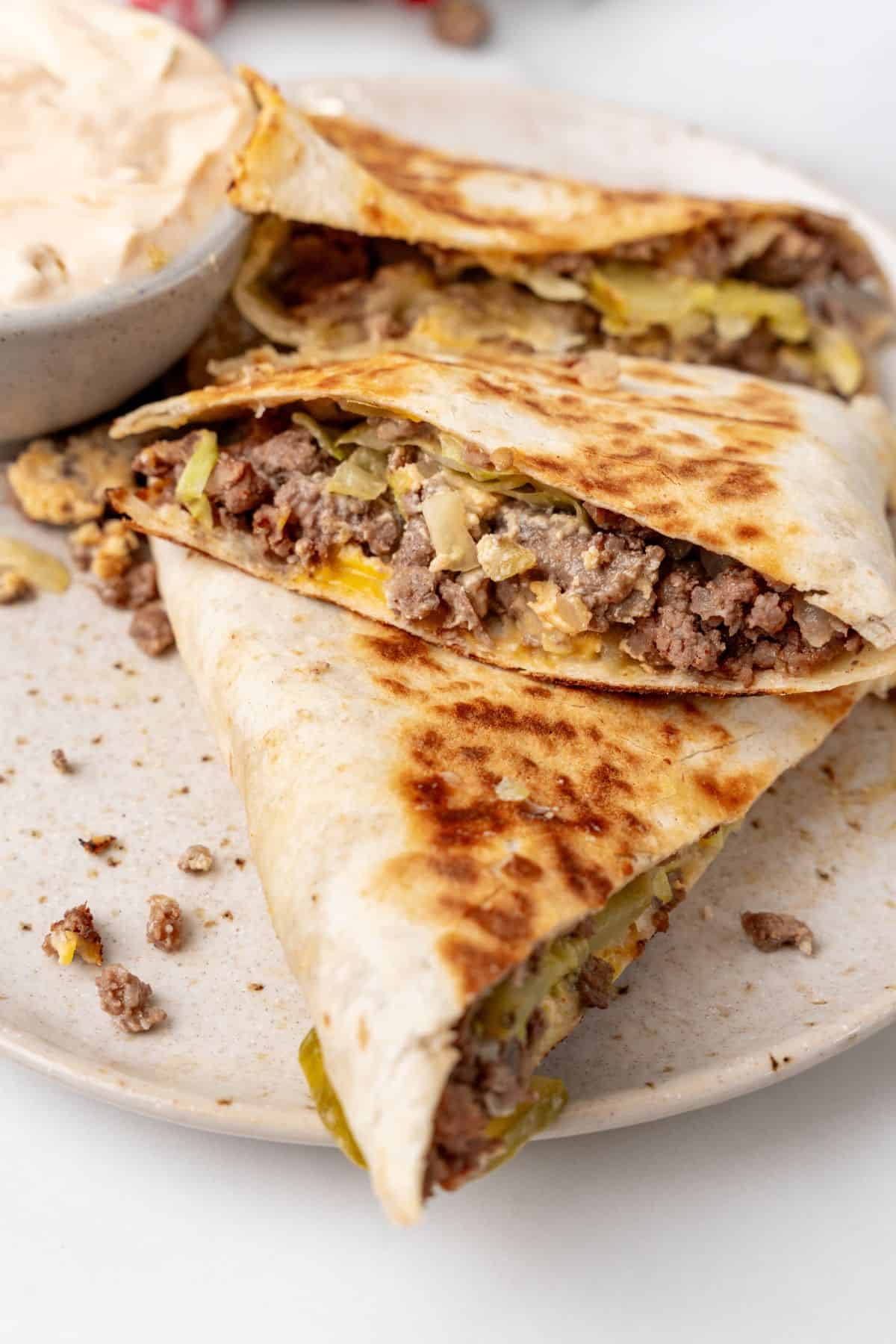 Cheesy Big Mac quesadillas with ground beef and melted cheese are stacked on a plate, accompanied by a bowl of dipping sauce.