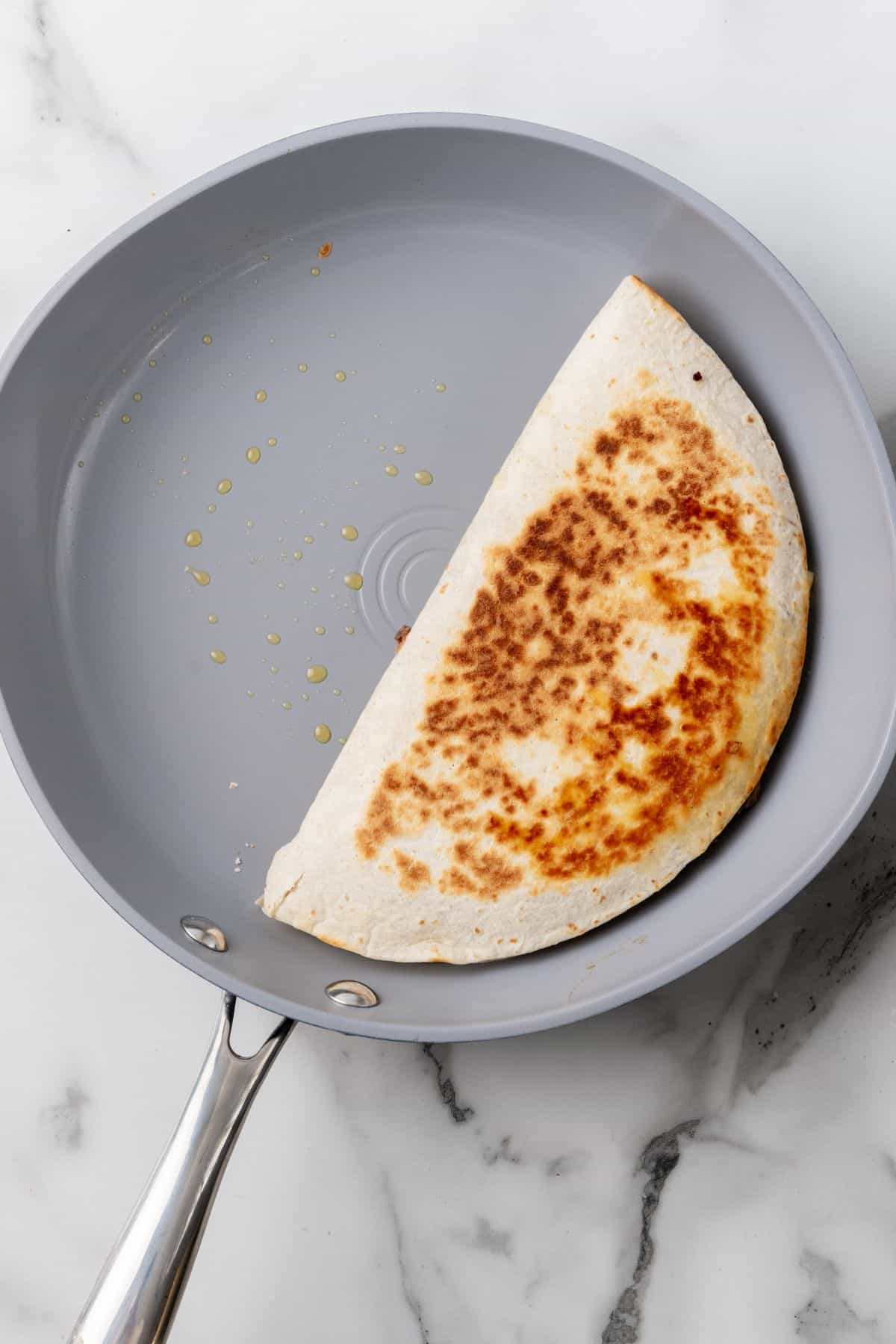 A folded quesadilla sizzles in a non-stick frying pan, reminiscent of a Big Mac's signature sizzle, with a few drops of oil glistening on the pan's surface.