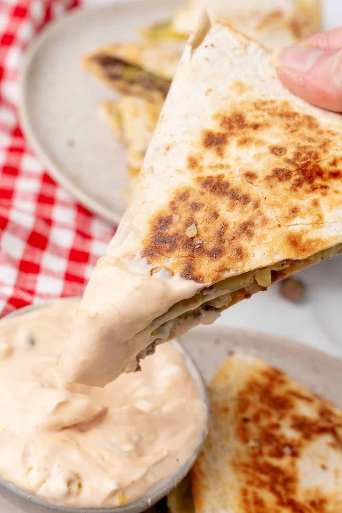 A hand dips a slice of quesadilla into a bowl of creamy dipping sauce, evoking the indulgence of a Big Mac, with a red and white checkered cloth in the background.