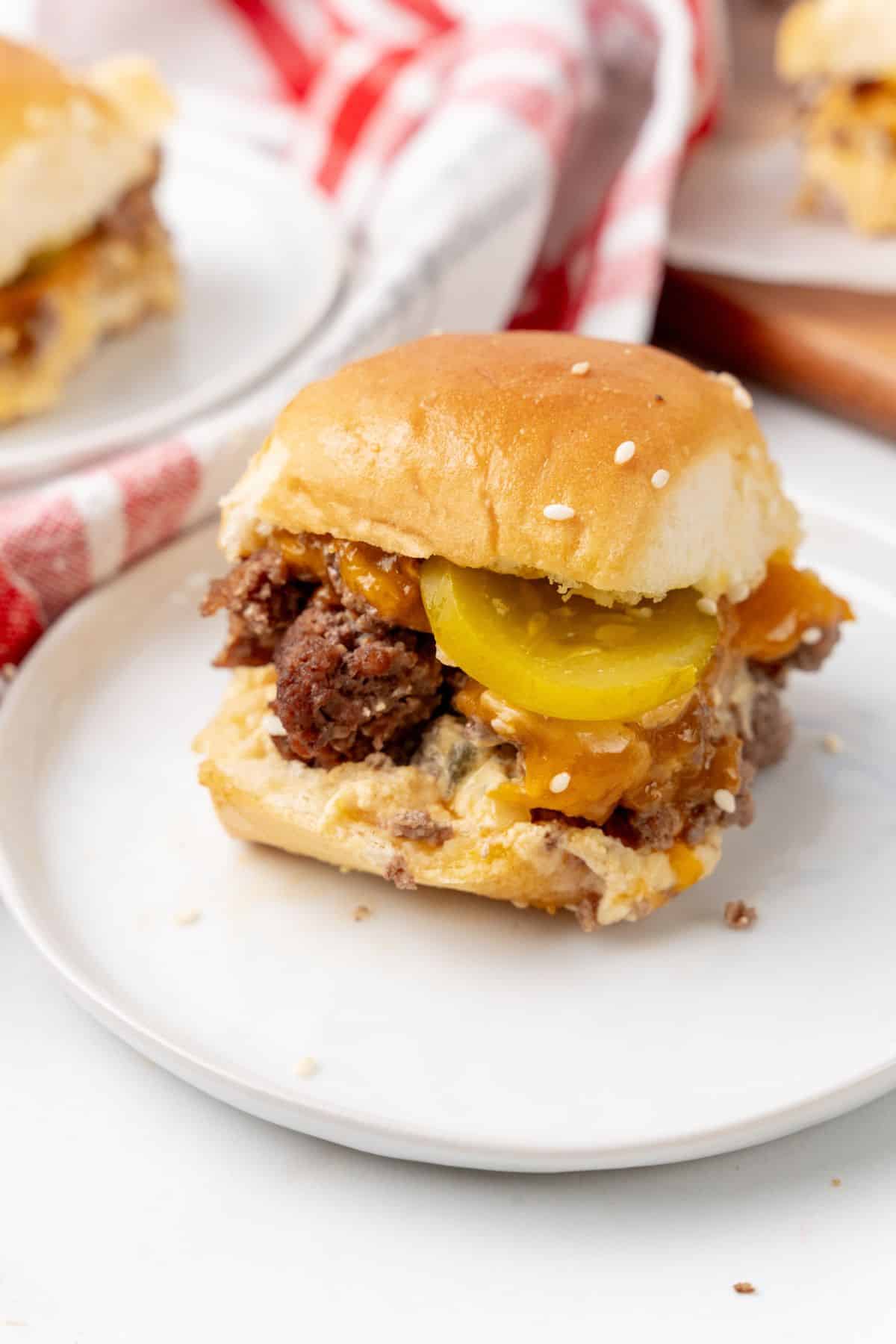 A Big Mac-inspired slider on a white plate, topped with pickles and cheese, sits on a table draped with a red and white checkered cloth in the background.