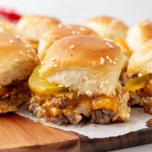 Close-up of several Big Mac Sliders on a wooden board. Each slider is topped with sesame seeds and contains beef, melted cheddar cheese, and pickles.