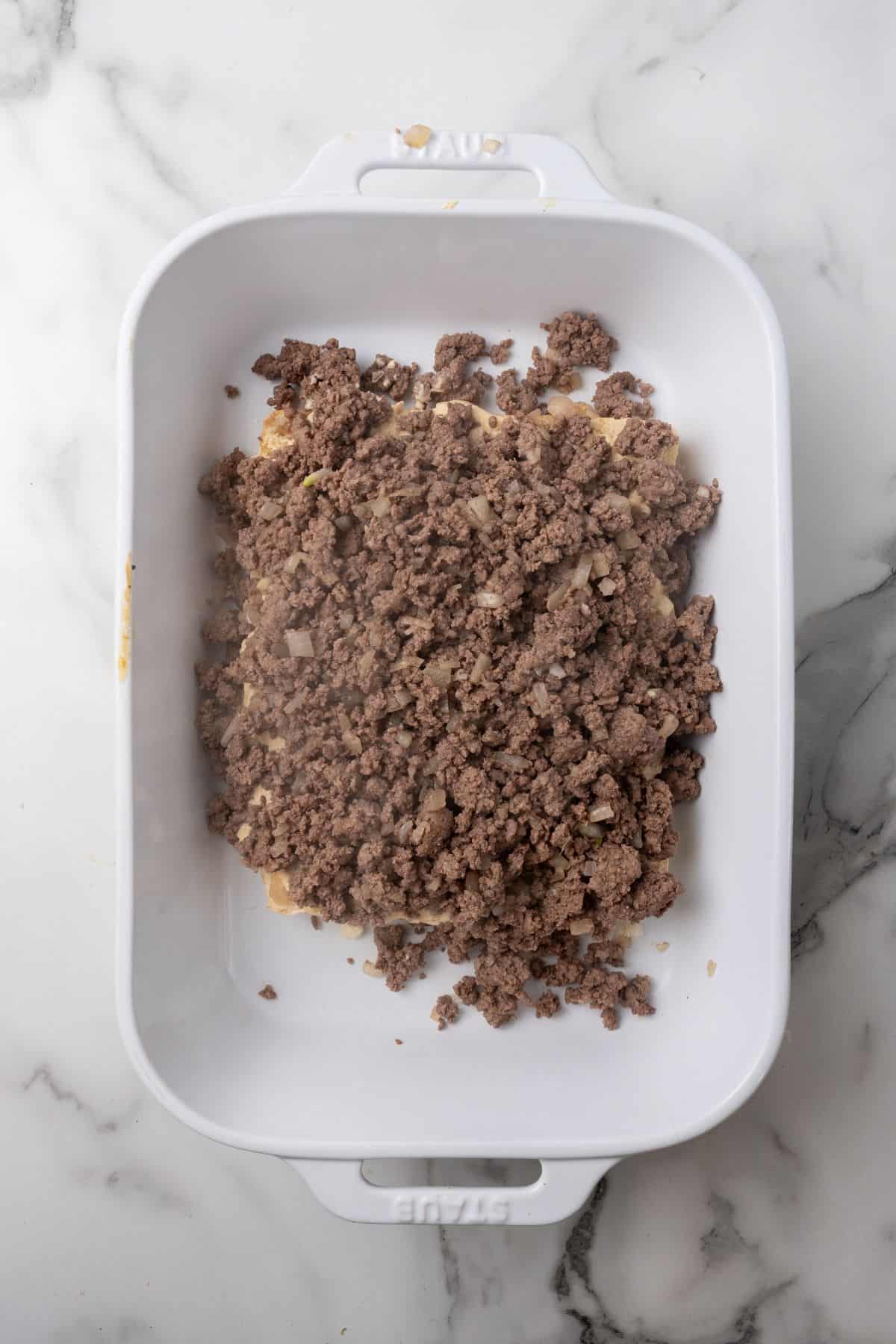 Ground beef mixed with sautéed onions in a white baking dish on a marble surface, creating the perfect base for delightful Big Mac sliders.