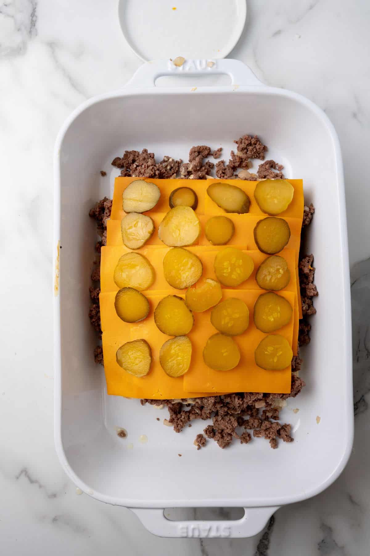Ground meat, reminiscent of Big Mac flavors, topped with cheddar cheese slices and pickles in a white baking dish on a marble surface.