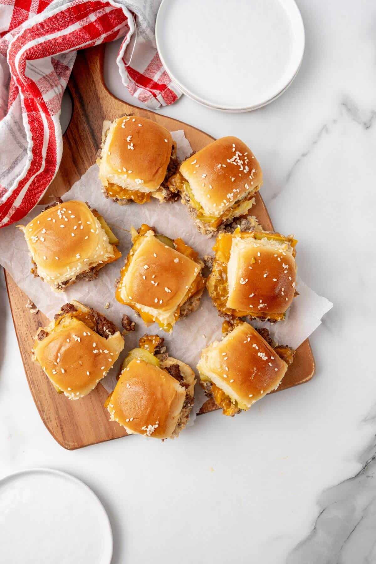 Mini sliders reminiscent of Big Mac flavors are topped with sesame seeds and arranged on a wooden board with parchment paper, accompanied by a red and white striped cloth. Nearby, two empty white plates await their delicious contents.