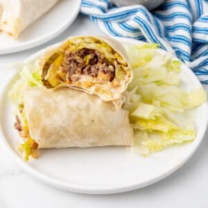 Burrito halves on a white plate resemble delicious Big Mac Wraps, filled with ground beef, lettuce, and cheese. A side of shredded lettuce complements the dish, with a striped cloth adding charm to the background.