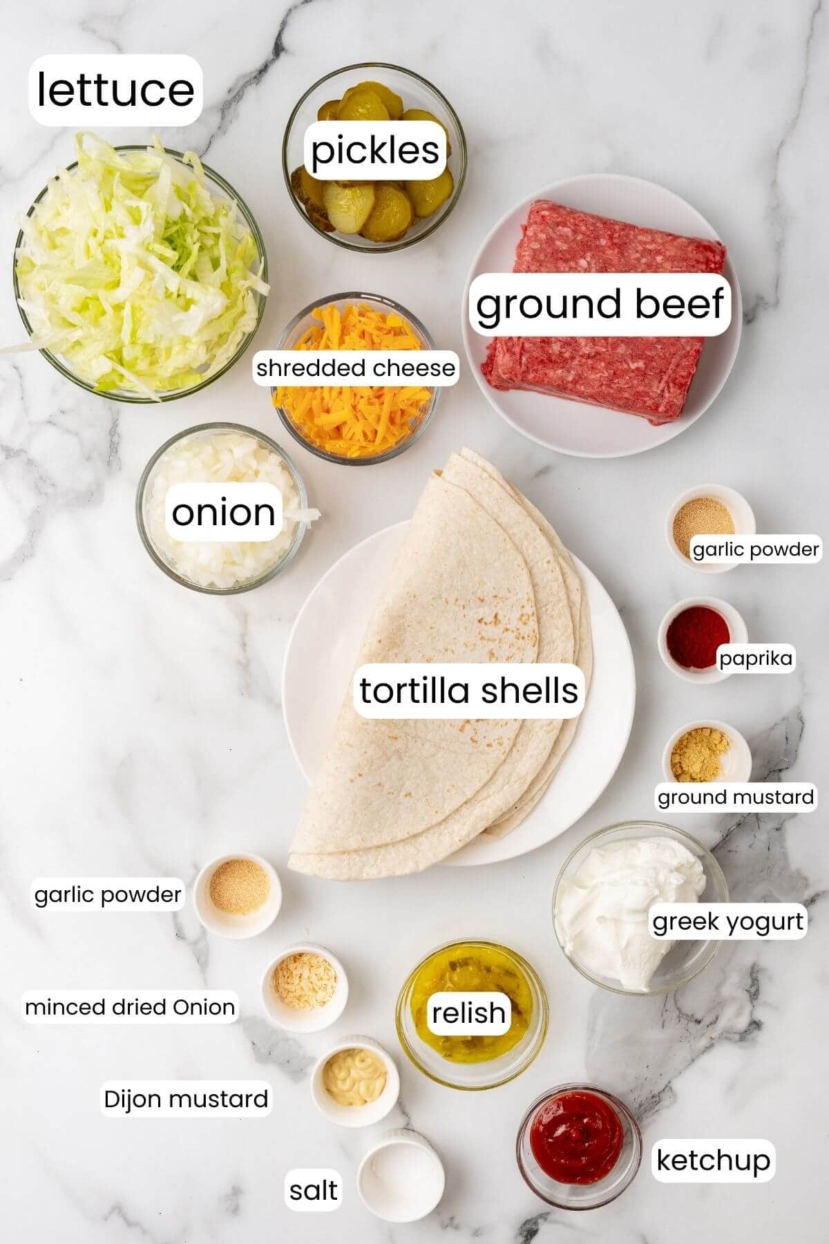 Ingredients for a recipe displayed on a marble surface: ground beef, tortilla shells, shredded cheese, lettuce, pickles, onion, yogurt, relish, ketchup, mustard, garlic powder and paprika. Create your own Big Mac wraps with these fresh spices and flavors.