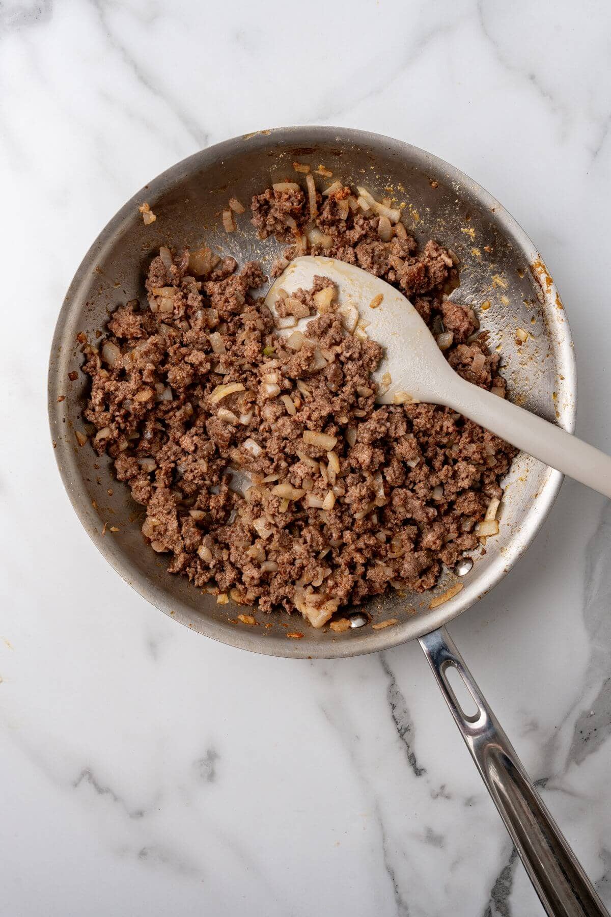 Ground beef and onions sizzle in a stainless steel skillet, expertly stirred with a white spatula on the marble surface. This savory blend channels the iconic flavors of Big Mac wraps, transforming every bite into a delectable experience.