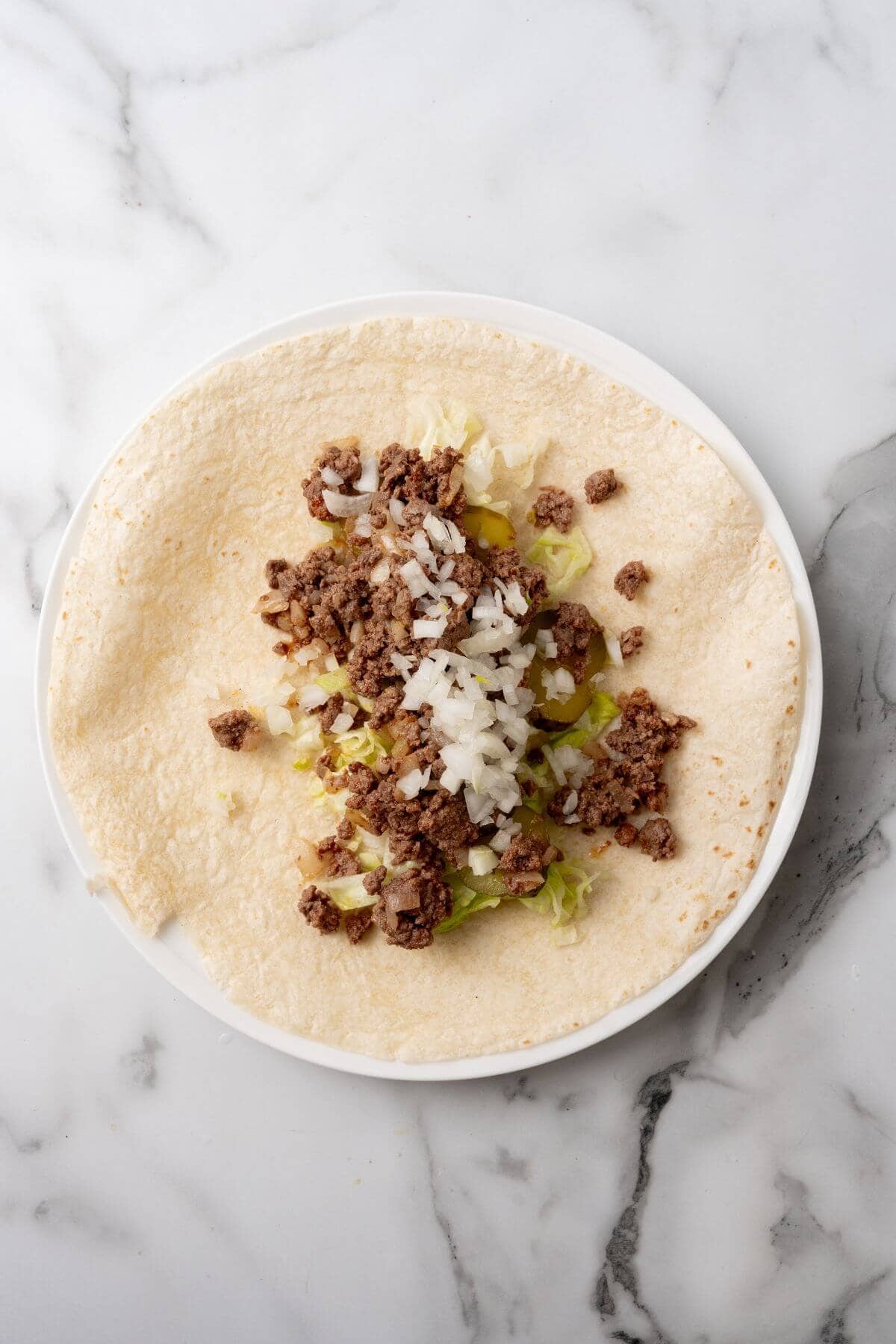 A tortilla wrap on a white plate with ground beef, chopped onions, and lettuce, reminiscent of a deconstructed Big Mac, sits elegantly on the marble surface.