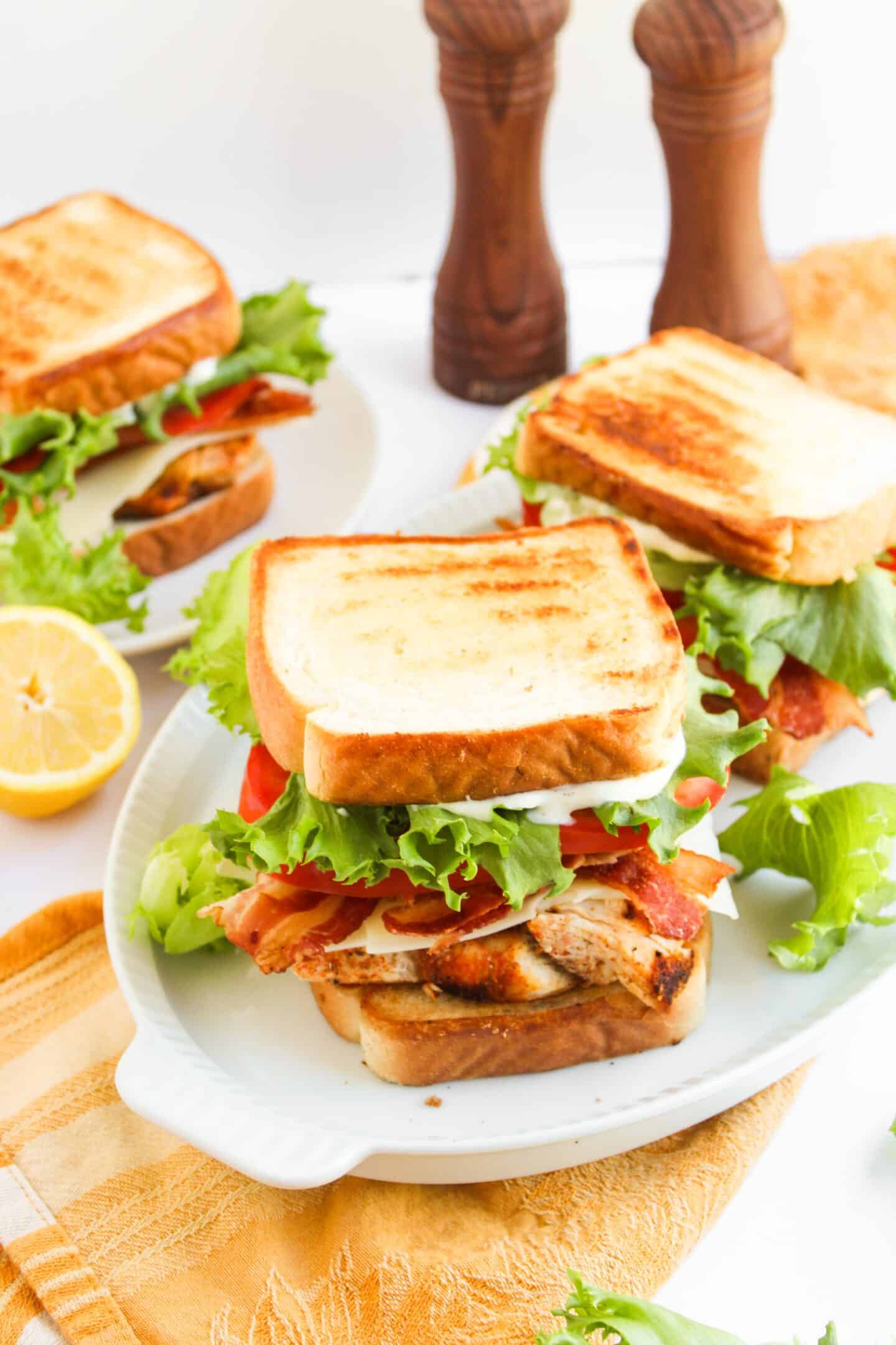 Three Blackened Chicken Club sandwiches with lettuce, tomato, and bacon are served on white plates, with a sliced lemon and two pepper mills in the background.