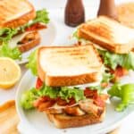 Three Blackened Chicken Club sandwiches with lettuce, tomato, and bacon are served on white plates, with a sliced lemon and two pepper mills in the background.