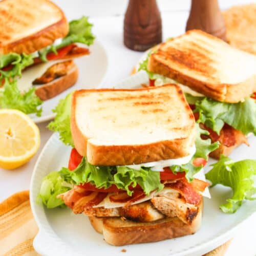 Three Blackened Chicken Club sandwiches with lettuce, tomato, and bacon are served on white plates, with a sliced lemon and two pepper mills in the background.