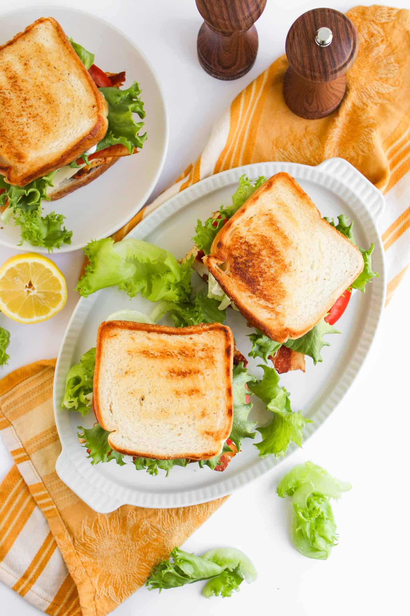 Two toasted Blackened Chicken Club sandwiches with lettuce on a white platter, accompanied by a lemon slice, a yellow-striped towel, and two wooden salt and pepper shakers.
