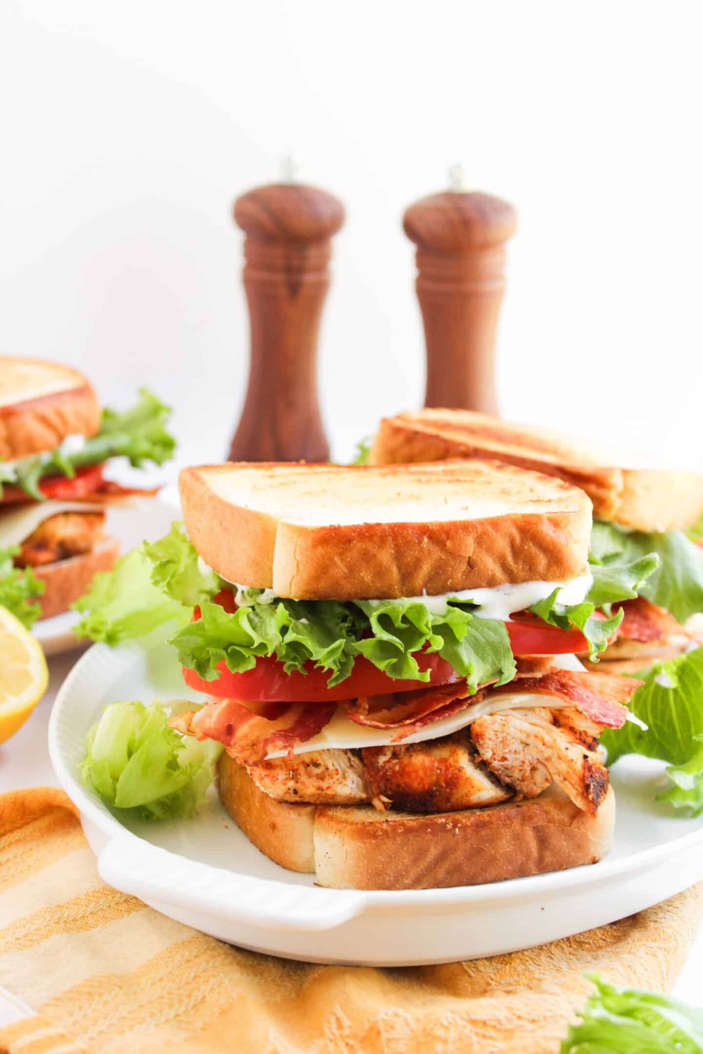 A Blackened Chicken Club sandwich with lettuce, tomato, bacon, and cheese on toasted bread is served on a white plate, with salt and pepper shakers in the background.