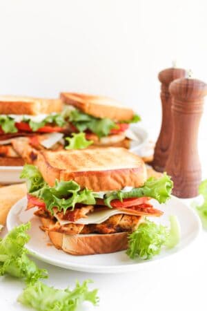 A toasted Blackened Chicken Club with lettuce, tomato, and cheese is on a white plate, with extra lettuce and two wooden pepper mills in the background.