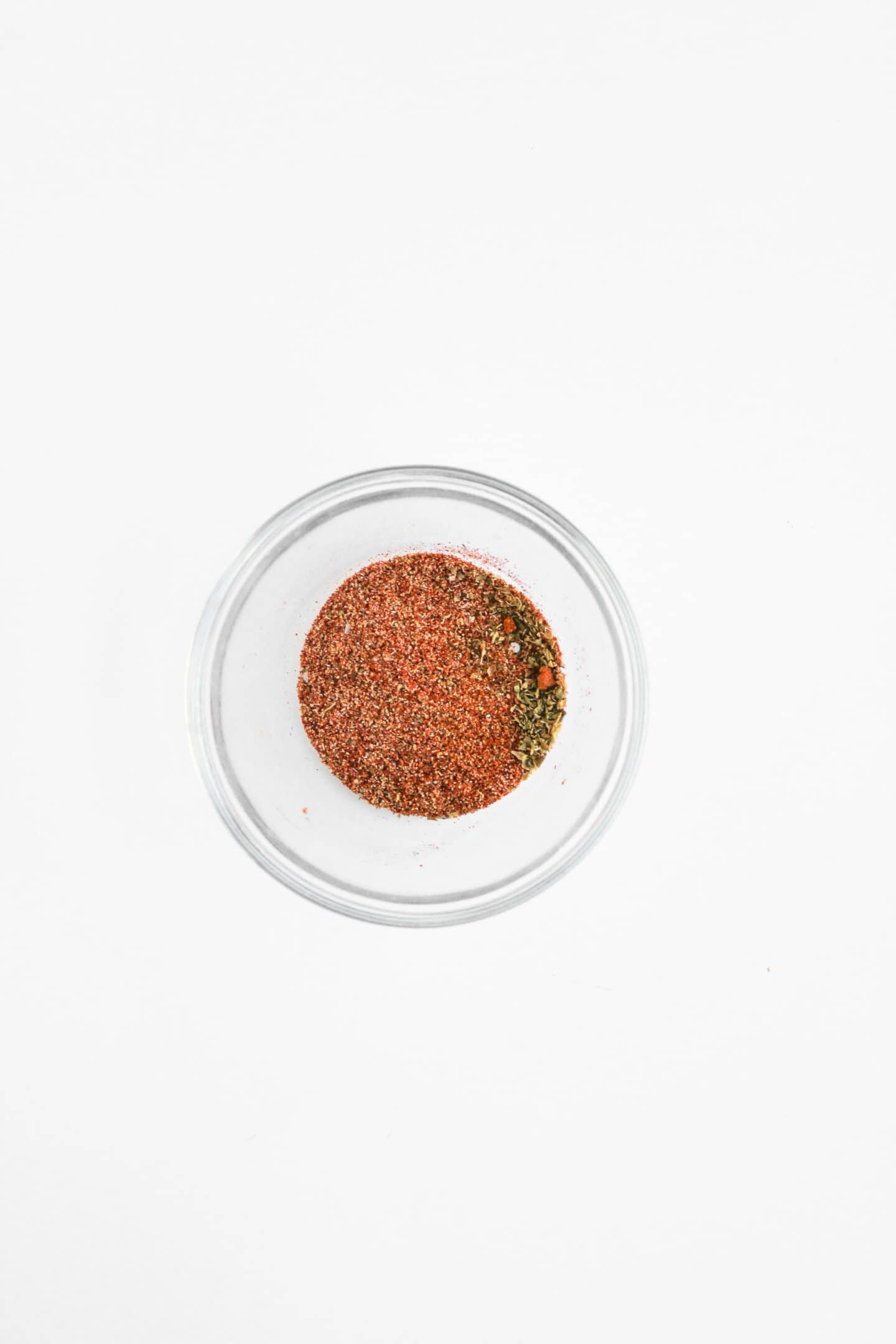 A small glass bowl containing a mixed red and green spice blend, perfect for seasoning Blackened Chicken Club sandwiches, viewed from above on a white background.