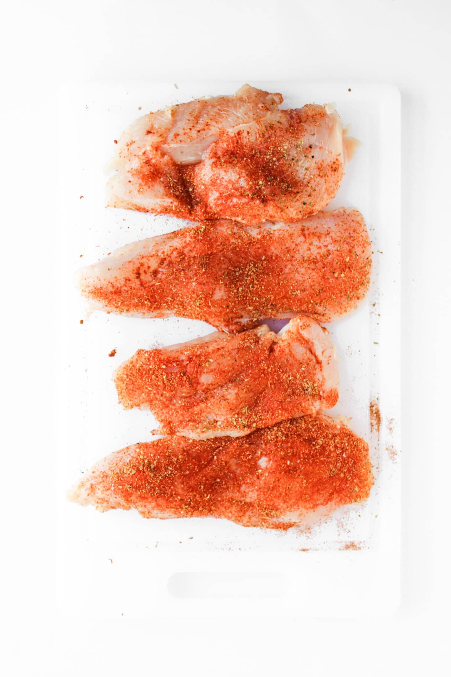 Four raw chicken breasts on a white cutting board, coated with a reddish spice blend for classic Blackened Chicken—perfect for hearty Blackened Chicken Club sandwiches.