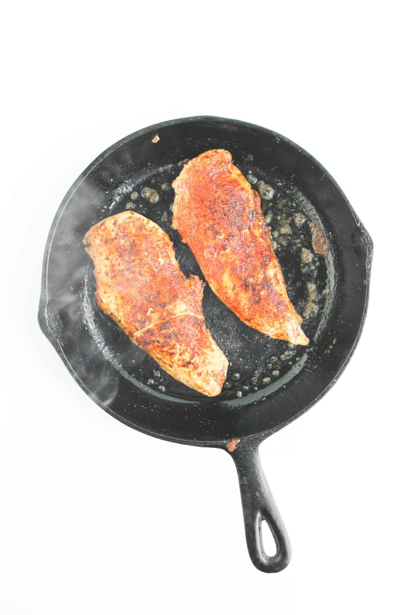 Two seasoned chicken breasts sizzle in a black cast iron skillet, with oil and steam rising—perfectly prepared for a flavorful Blackened Chicken Club.