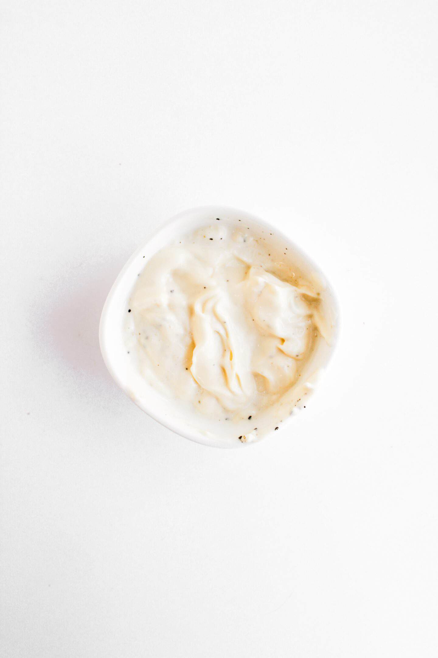 A small white bowl filled with creamy white sauce, lightly swirled and speckled with black pepper, perfect for drizzling over Blackened Chicken Clubs, sits on a white background.
