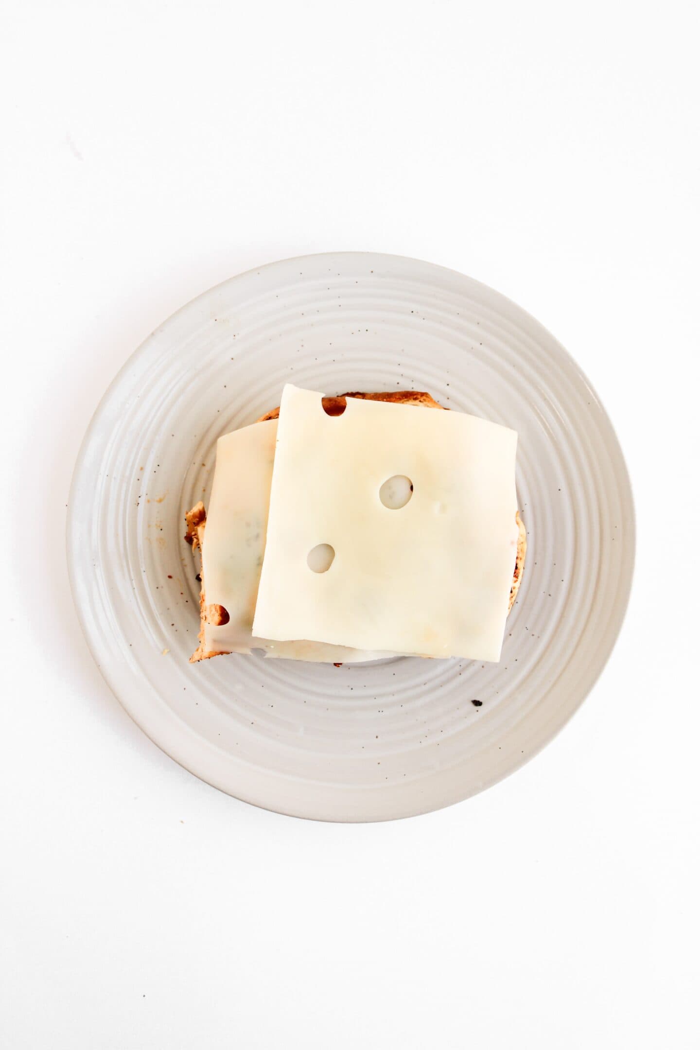 A slice of bread topped with two slices of Swiss cheese, reminiscent of classic Chicken Clubs, sits on a light gray plate and is viewed from above.