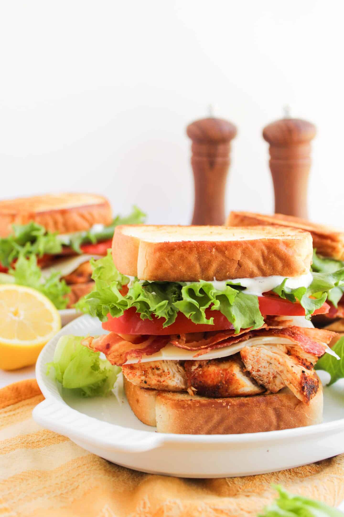 A Blackened Chicken Club sandwich with lettuce, tomato, bacon, and cheese on toasted bread sits on a white plate, with a lemon half and salt and pepper shakers in the background.
