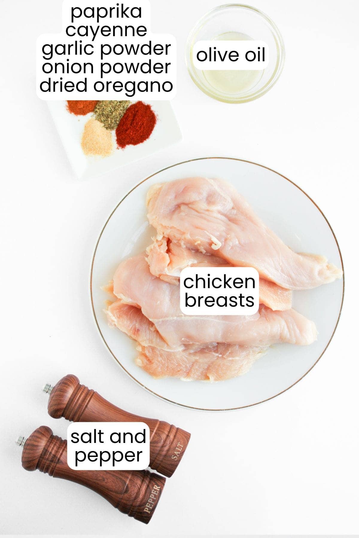 A plate with raw chicken breasts for Blackened Chicken Clubs, a bowl of olive oil, assorted spices, and salt and pepper grinders arranged on a white surface.