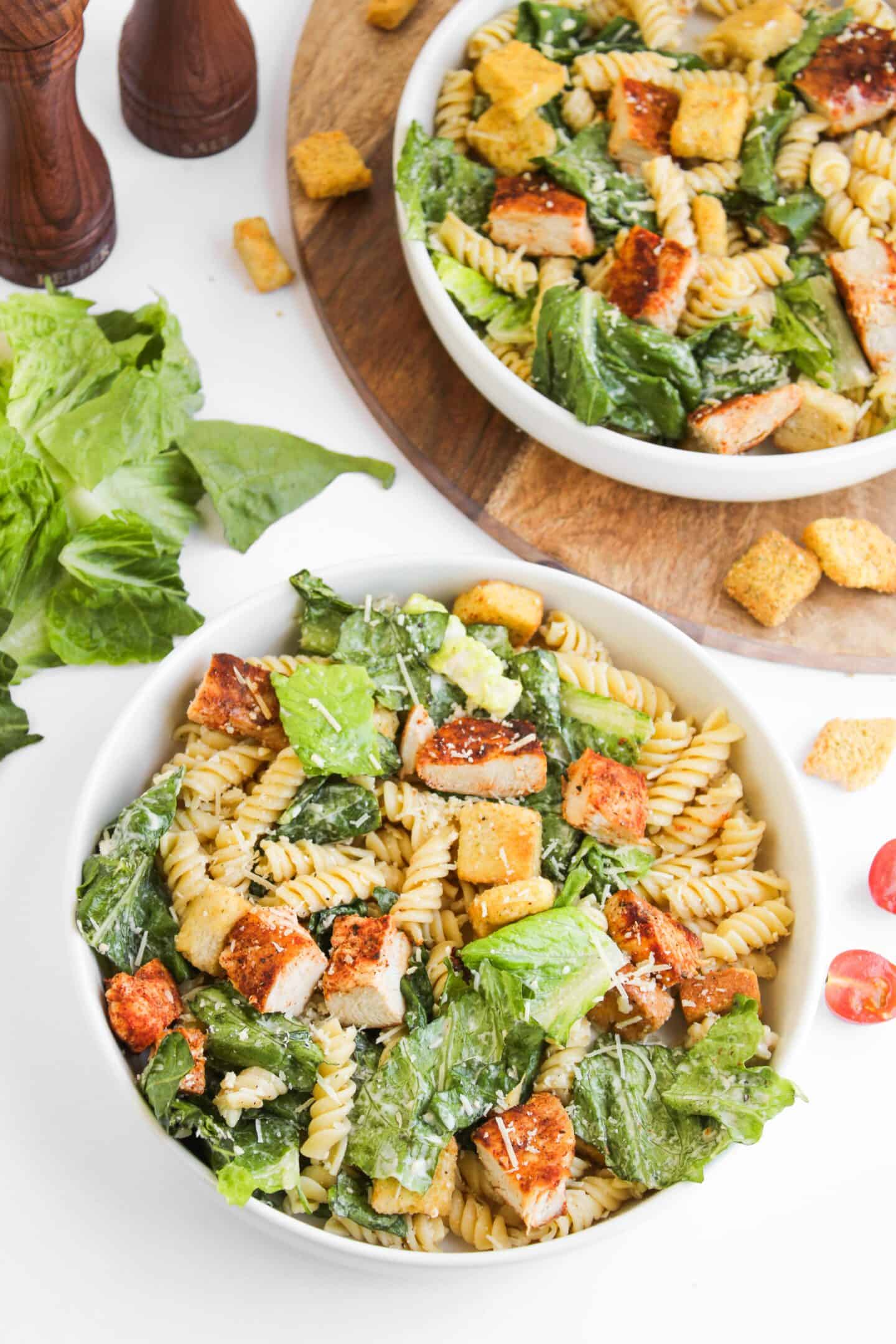 Two bowls of Chicken Caesar Pasta Salad with rotini, grilled chicken, romaine lettuce, croutons, and grated cheese on a white surface, ingredients scattered around for a fresh take on the classic pasta salad.