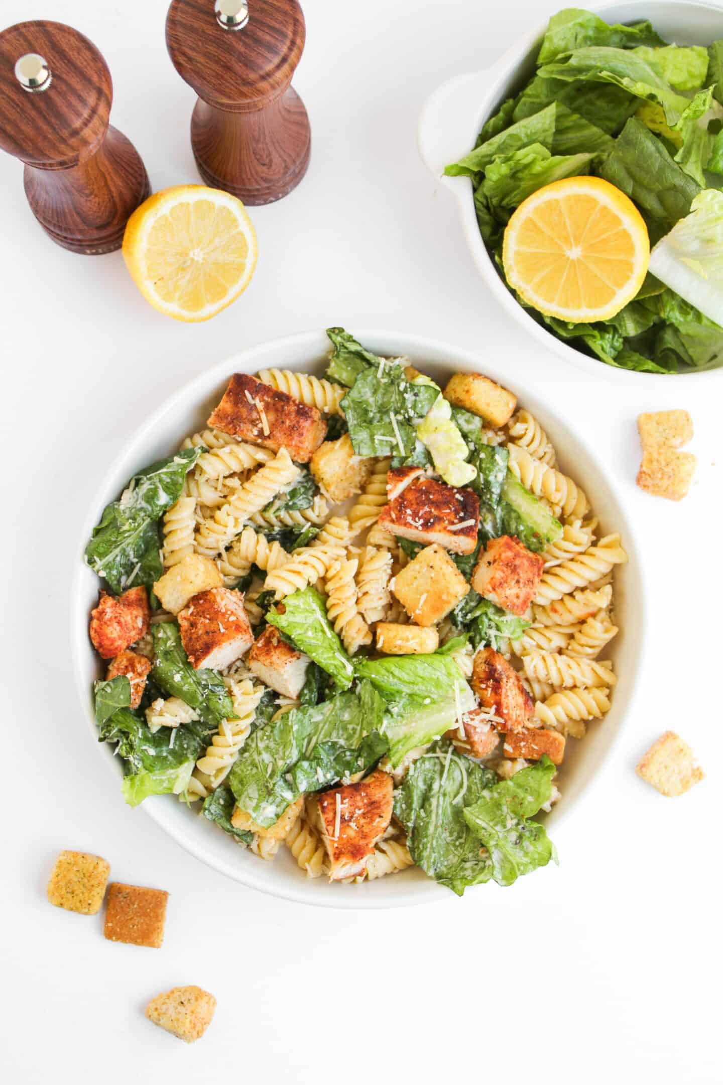 A bowl of Chicken Caesar Pasta Salad with rotini, romaine lettuce, croutons, and Parmesan cheese sits next to a halved lemon, salad bowl, and wooden salt and pepper shakers.