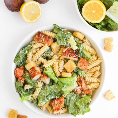 A bowl of Chicken Caesar Pasta Salad with rotini, romaine lettuce, croutons, and Parmesan cheese sits next to a halved lemon, salad bowl, and wooden salt and pepper shakers.