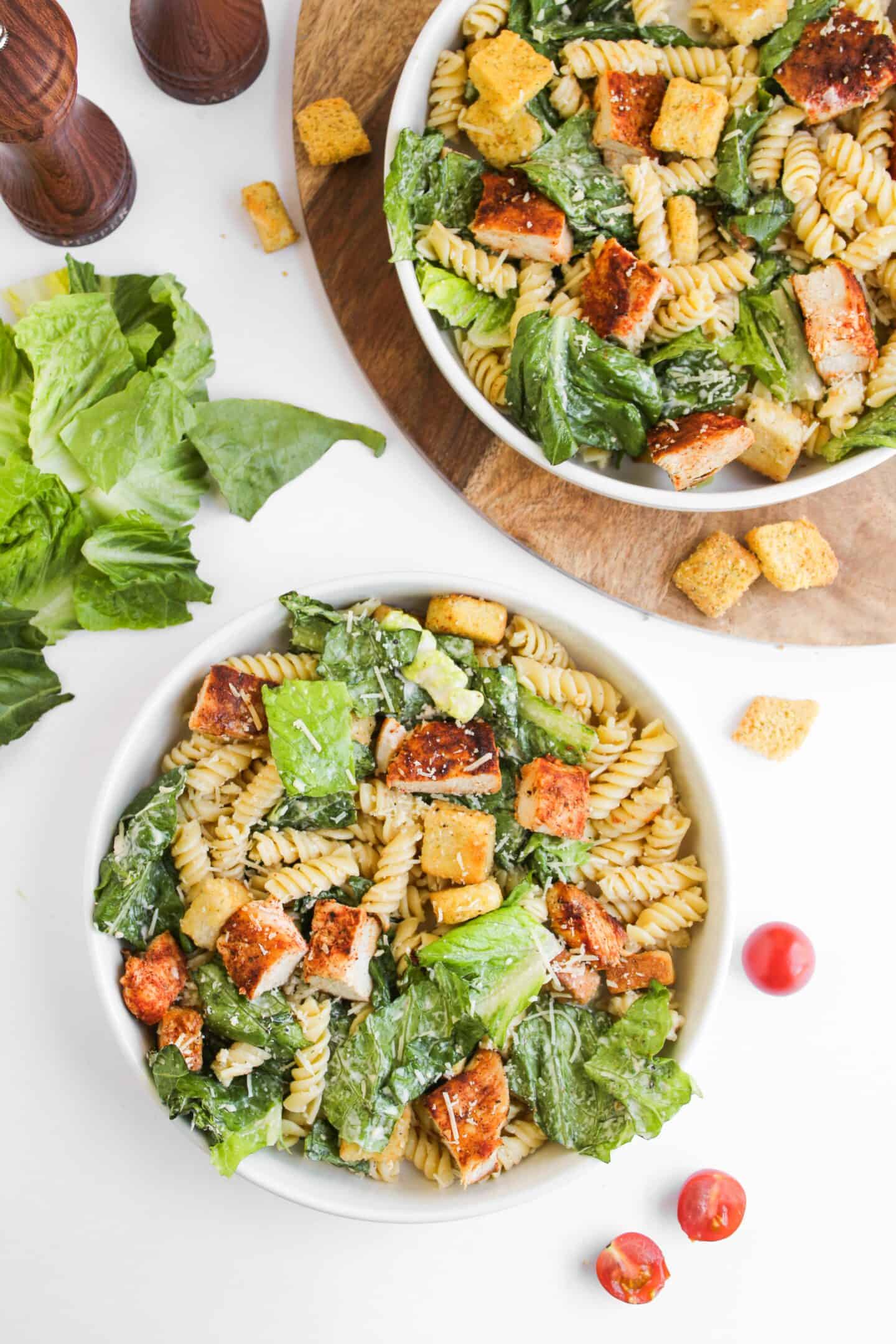 Two bowls of Chicken Caesar Pasta Salad with rotini, grilled chicken, romaine lettuce, croutons, and Caesar dressing on a white surface with loose lettuce, croutons, and cherry tomatoes.