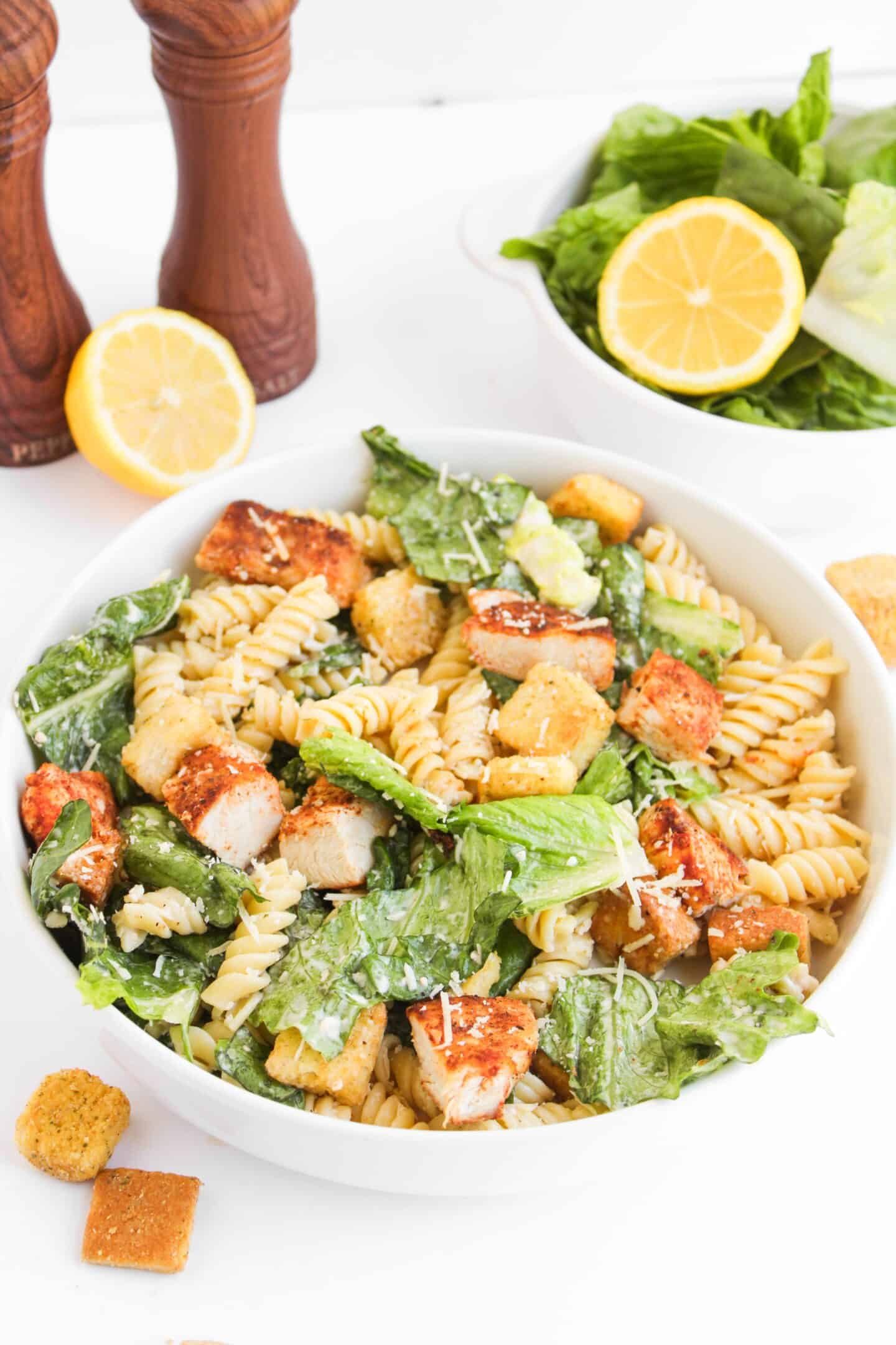 A bowl of Chicken Caesar Pasta Salad with rotini, romaine lettuce, grilled chicken, croutons, and grated cheese. In the background, a fresh salad with lemon slices and pepper mills complete this Chicken Caesar-inspired meal.