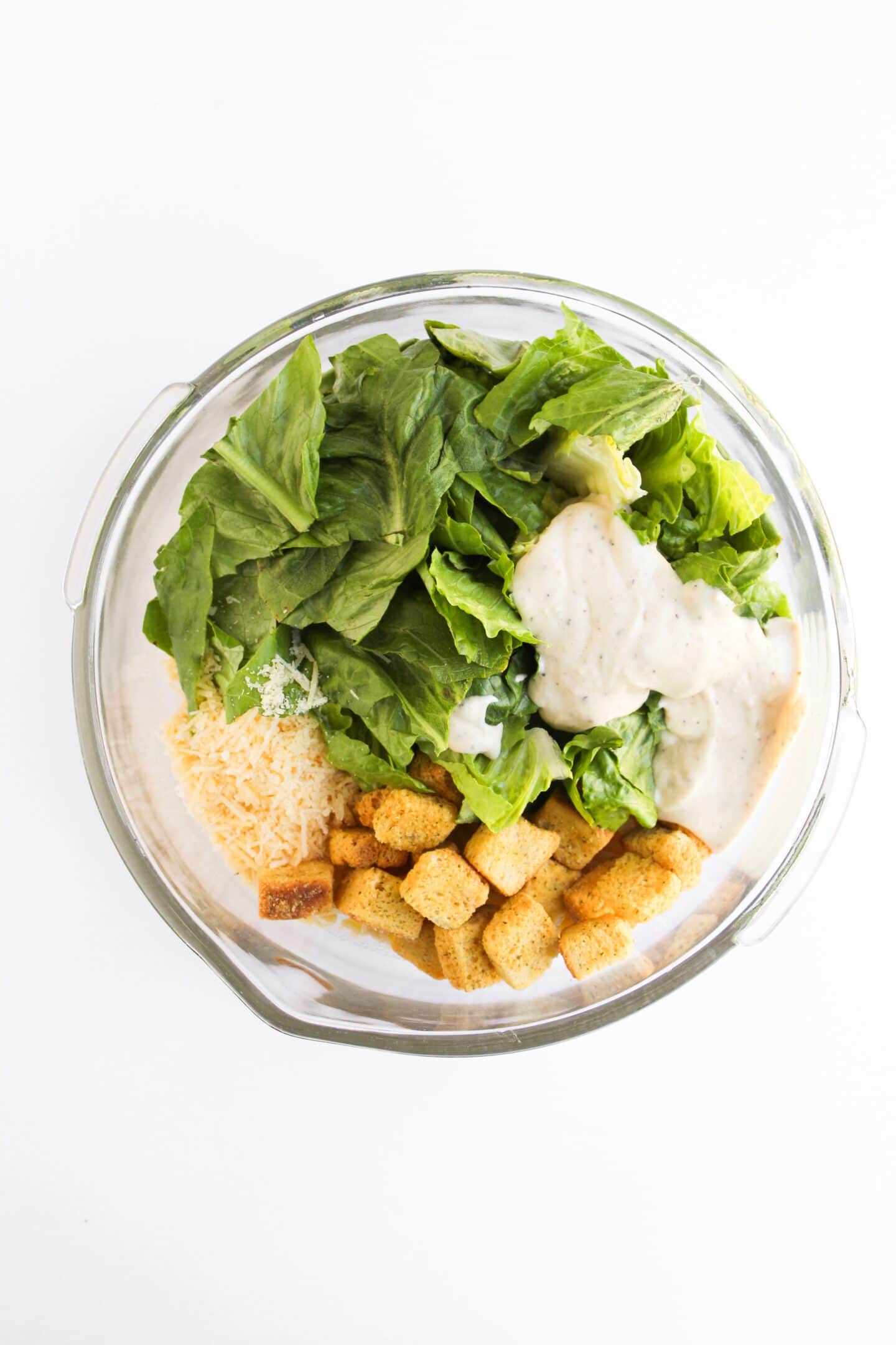 A glass bowl containing romaine lettuce, Chicken Caesar dressing, shredded Parmesan cheese, croutons, and pasta on a white background for a delicious Pasta Salad.