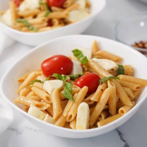 Tuscan bowls of penne pasta salad with cherry tomatoes, mozzarella, and fresh basil leaves rest invitingly on a marble surface, offering a healthy taste of Italy.