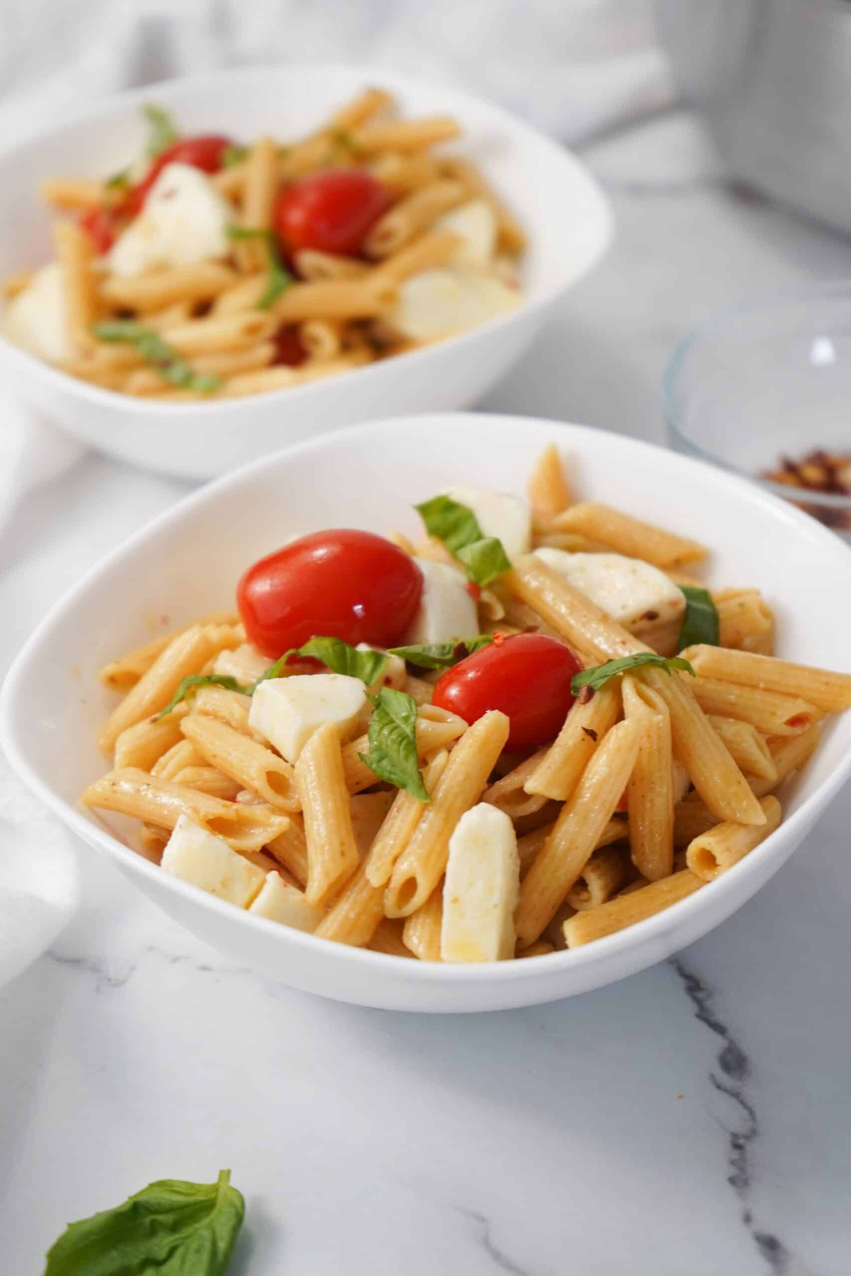 Tuscan bowls of penne pasta salad with cherry tomatoes, mozzarella, and fresh basil leaves rest invitingly on a marble surface, offering a healthy taste of Italy.
