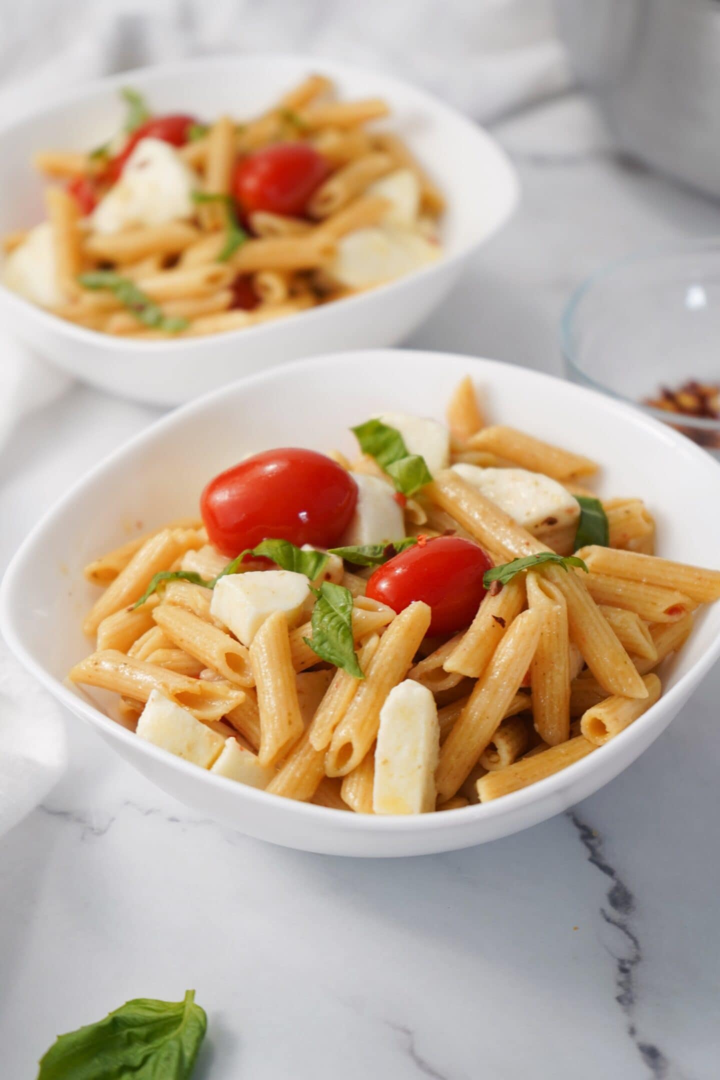 Two bowls of healthy penne pasta salad with cherry tomatoes, mozzarella, and fresh basil sit elegantly on a white marble surface, evoking the flavors of a Tuscan summer.