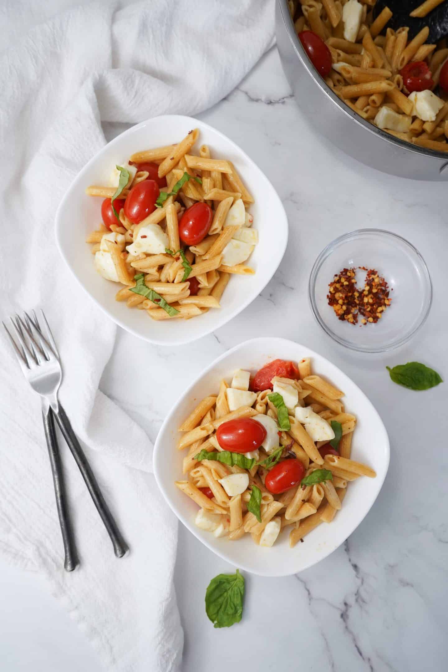 Two bowls of healthy Tuscan penne pasta with cherry tomatoes, mozzarella, and basil sit elegantly on a marble surface. Nearby, a pot of pasta and a small bowl of chili flakes await, accompanied by forks and a napkin for a delightful dining experience.