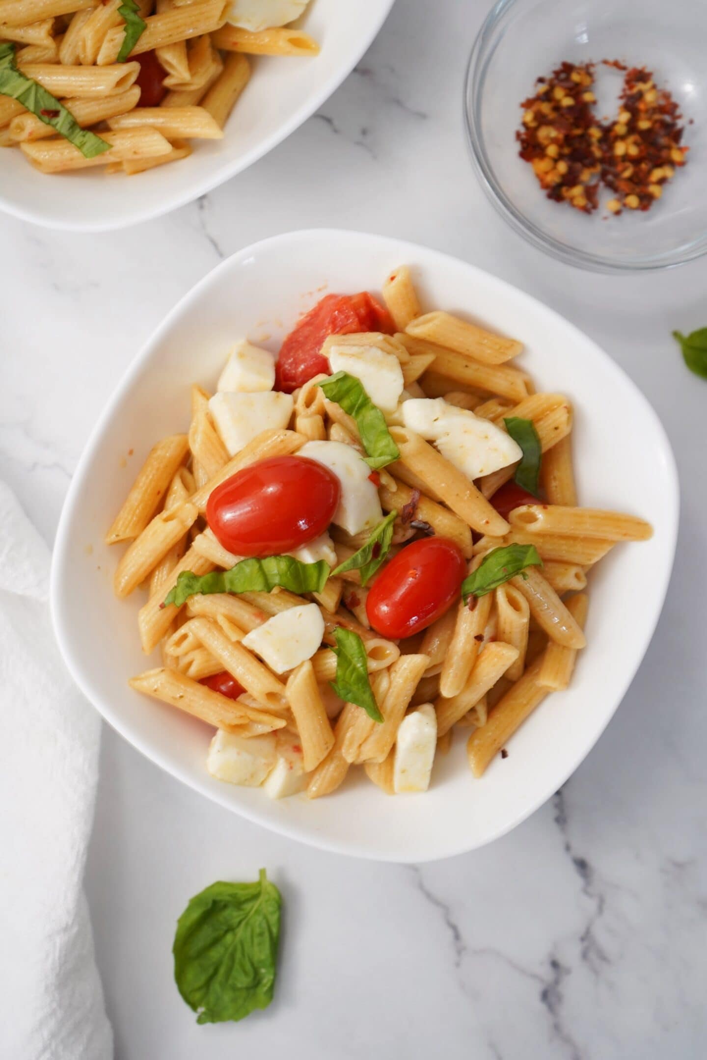 A bowl of Tuscan penne pasta with cherry tomatoes, mozzarella, and basil rests on a white marble surface. A small dish with red pepper flakes is nearby, adding a healthy touch of spice.
