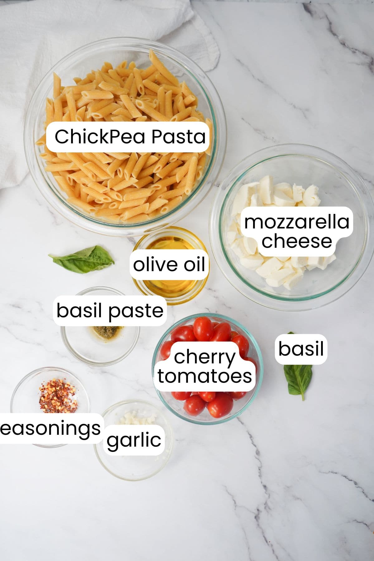 Ingredients for a healthy Tuscan pasta dish arranged on a marble surface: Chickpea pasta, mozzarella cheese, olive oil, basil paste, cherry tomatoes, basil, seasonings, and garlic.