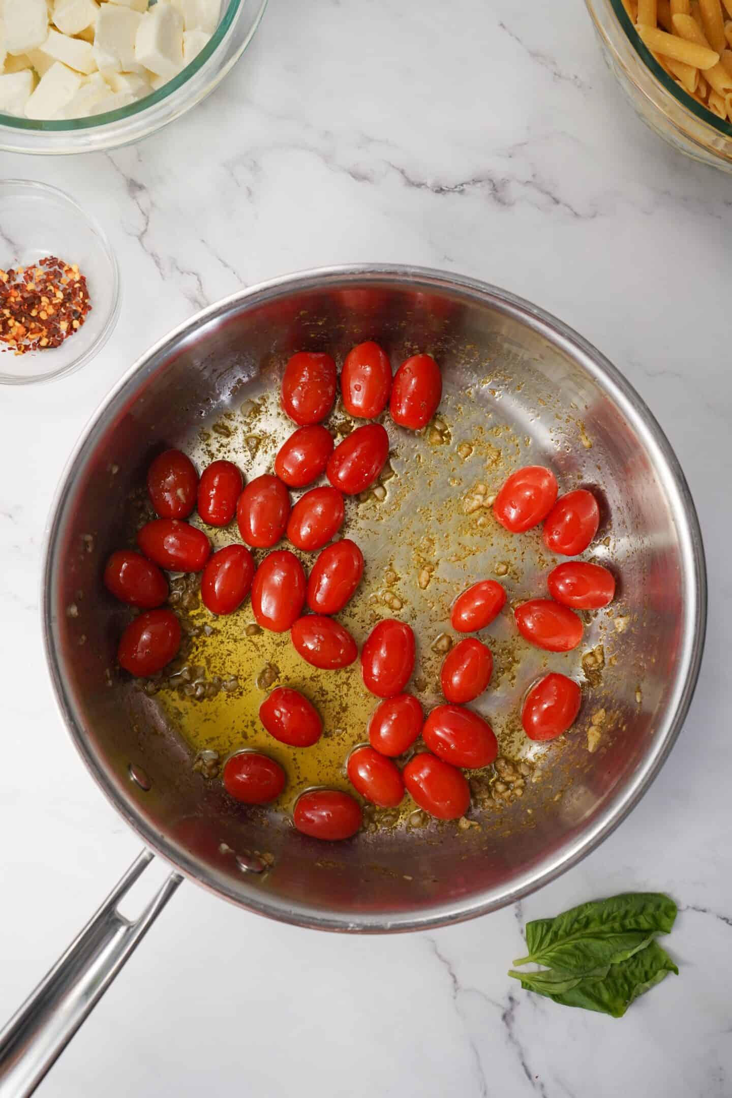 Cherry tomatoes sizzle in a Tuscan pan with olive oil and garlic. Nearby, bowls of healthy pasta, mozzarella, and red pepper flakes rest invitingly on the marble surface.