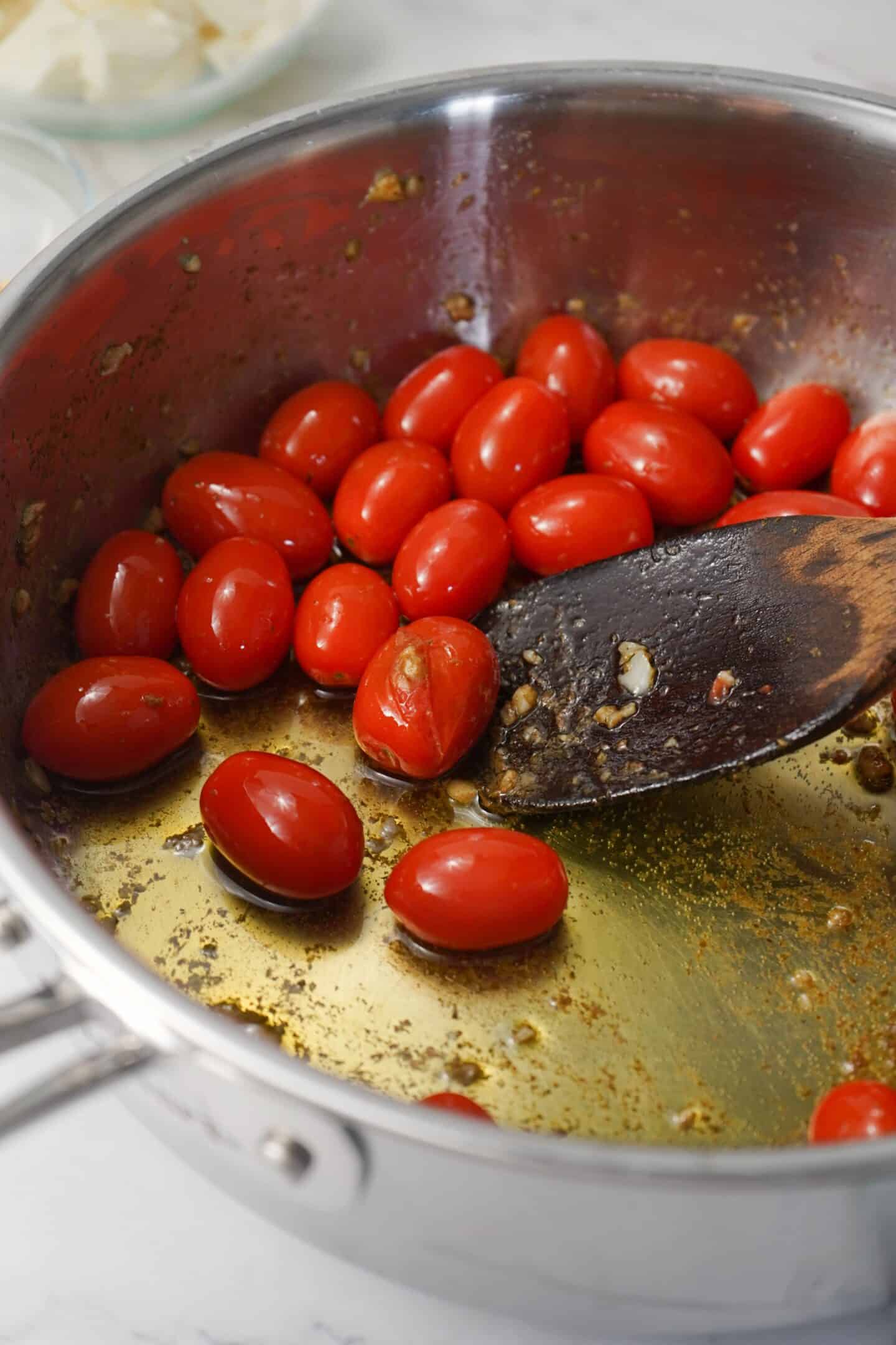 Tuscan cherry tomatoes sizzle in a stainless steel pan with oil and seasonings, stirred with a wooden spoon to create a healthy and delicious accompaniment for pasta.