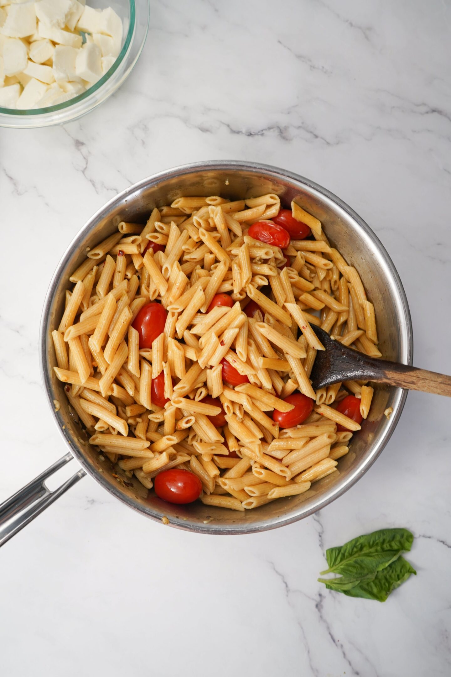 A Tuscan pot of penne pasta with cherry tomatoes and a wooden spoon rests on a marble surface. Nearby, a bowl of cheese and fragrant basil leaves completes this healthy culinary scene.